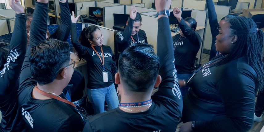 A group of people in long-sleeved, dark J. Crew t-shirts all hold their right hands in the air in a cheer or in agreement. They are standing in the center of a group of cubicles in an office.