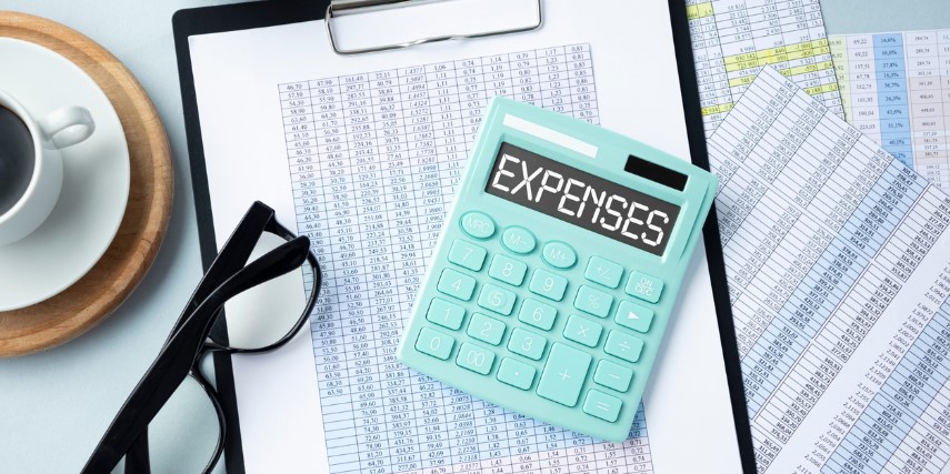 A clipboard holds a printed spreadsheet with lists of figures. A calculator and a pair of eyeglasses also rest on the clipboard with a cup of coffee next to it.