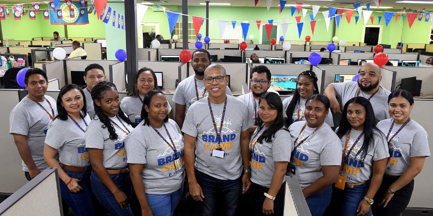 A group of approximately 15 people stand together smiling. They are all dressed in a Protel BPO t-shirt that says, "Brand Camp."